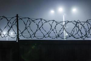 Silhouette von Stacheldraht auf einem Zaun vor dem Hintergrund verschwommener Laternenpfähle in nebliger Nacht foto