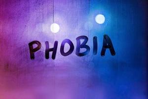 das Wort Phobie handschriftlich auf nasser, nebliger Fensterglasoberfläche mit lila-blauem Hintergrundlicht foto