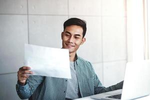 glücklicher junger asiatischer geschäftsmann, der an seinem arbeitsplatz an einem computer-laptop arbeitet. lächelnd und Papierarbeitsblatt betrachtend foto