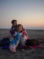Liebendes junges Paar, das am Strand neben dem Lagerfeuer sitzt und Bier trinkt foto
