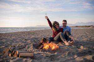 junges Paar sitzt am Strand neben Lagerfeuer und trinkt Bier foto
