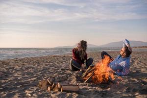 junges Paar sitzt am Strand neben Lagerfeuer und trinkt Bier foto