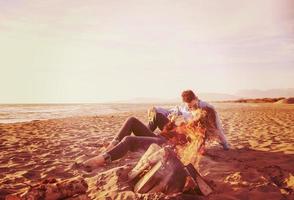 junges Paar sitzt am Strand neben Lagerfeuer und trinkt Bier foto