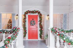 idee für die dekoration der weihnachtsterrasse. Hauseingang mit roter Tür für den Urlaub dekoriert. rote und grüne Kranzgirlande aus Tannenzweigen und Lichtern am Geländer. Heiligabend zu Hause foto