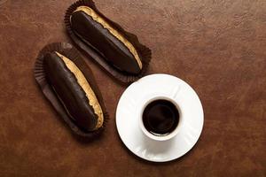 Kaffee, Schokoladen-Eclair, Kaffee in einer weißen Tasse, weiße Untertasse, auf einem braunen Tisch, Eclair auf Papierständer foto