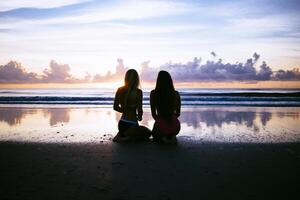 eine schwache Aufnahme von zwei Frauen im Bikini, die an einem nassen Strand knien und den Sonnenuntergang bewundern foto