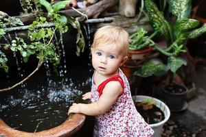 Kleines Mädchen mit blonden Haaren und blauen Augen im Sommerkleid steht im Garten mit Topfpflanzen und Wasserfall. foto