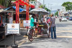 georgetown, penang, malaysia, 13. februar 2020. alter mann in der nähe von trishaw auf einer belebten straße der stadt mit autos, cafés und geschäften. foto