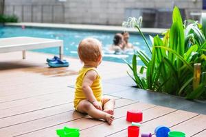 Kleines Mädchen mit schmutzigen Knien, das draußen mit buntem Plastikbauer spielt und auf den Swimmingpool schaut. foto