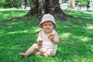 entzückendes babymädchen in weißer kleidung und panama spielt mit trockenen gelben blättern auf gras. sonniger sommertag im park. foto