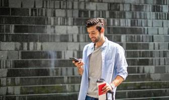 Junger kaukasischer Mann mit Smartphone, der die Kaffeetasse hält und den Koffer zieht und auf seinen Freund wartet, um gemeinsam zu reisen. foto