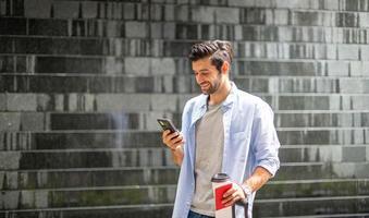 Junger kaukasischer Mann mit Smartphone, der die Kaffeetasse hält und den Koffer zieht und auf seinen Freund wartet, um gemeinsam zu reisen. foto