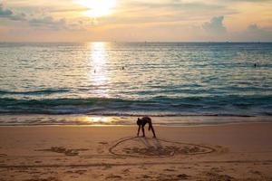 Mädchen, das Herz auf Sand am Strand nahe dem Meer im Sonnenuntergang malt foto