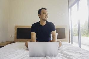 glücklicher junger mann, der vom bett aus arbeitet. Stell den Laptop aufs Bett und tippe auf der Tastatur. foto