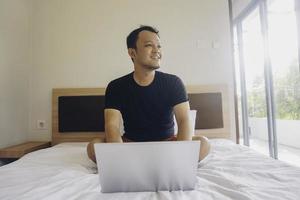 glücklicher junger mann, der vom bett aus arbeitet. Stell den Laptop aufs Bett und tippe auf der Tastatur. foto