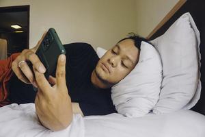 müder junger mann, der sein telefon überprüft, während er im bett liegt foto