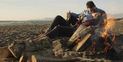 Liebendes junges Paar, das am Strand neben dem Lagerfeuer sitzt und Bier trinkt foto