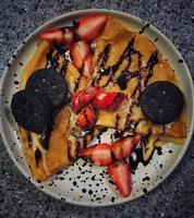 Leckere und appetitliche Pfannkuchen mit Erdbeeren und Keksen in Schokoladen- und Vanillecreme auf einem Teller mit schwarzen Punkten foto