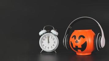 Halloween-Kürbis aus Kunststoff mit Kopfhörern und weißem Vintage-Wecker um 12 Uhr, isoliert auf schwarzem Hintergrund mit Kopierraum. Halloween-Musik oder Podcast. foto