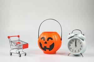 halloween-kürbiseimer mit einkaufswagen und weißem vintage-wecker 12 uhr. getrennt auf weißem Hintergrund mit Kopienraum. Halloween-Feiertags-Shopping-Konzept um Mitternacht. foto