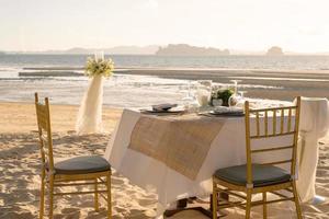 Schöner Tisch für ein romantisches Abendessen am Strand mit Blumen und Kerzen. Catering für ein romantisches Date, eine Hochzeit oder einen Flitterwochenhintergrund. Abendessen am Strand bei Sonnenuntergang. ausgewählten Fokus. foto