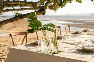 Schöner Tisch für ein romantisches Abendessen am Strand mit Blumen und Kerzen. Catering für ein romantisches Date, eine Hochzeit oder einen Flitterwochenhintergrund. Abendessen am Strand bei Sonnenuntergang. ausgewählten Fokus. foto