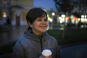 Ein Teenager mit positiven Emotionen und einem Pappbecher in der Hand auf der Straße an einem dunklen Herbstabend vor dem Hintergrund der Lichter der Stadt. foto