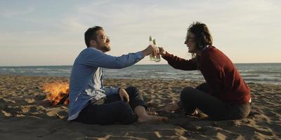 Liebendes junges Paar, das am Strand neben dem Lagerfeuer sitzt und Bier trinkt