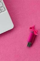 Eine leuchtend rosa USB-Flash-Speicherkarte mit einer rosa Schleife liegt auf einer Decke aus weichem und pelzigem hellrosa Fleece-Stoff neben einem weißen Laptop. klassisches weibliches Geschenkdesign für eine Speicherkarte foto