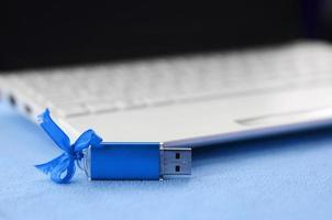 Eine leuchtend blaue USB-Flash-Speicherkarte mit einer blauen Schleife liegt auf einer Decke aus weichem, pelzigem hellblauem Fleece-Stoff neben einem weißen Laptop. klassisches weibliches Geschenkdesign für eine Speicherkarte foto