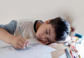 Kind, das auf Papier zeichnet, Porträtjunge, der mit dem Kopf nach unten auf der Hand liegt, während er graue Stiftmalerei verwendet, Kind mit denkendem Gesicht, das Kunst und Handwerk für Hausaufgaben macht. foto