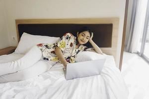 eine junge lächelnde Frau, die mit ihrem Laptop arbeitet, während sie auf dem Bett liegt. foto