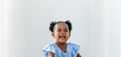 Afroamerikanisches kleines Mädchen, das ein süßes blaues Kleid mit lockigem Haar trägt, glückliches Lächeln auf weißem Hintergrund. glückliche kinder zu hause foto