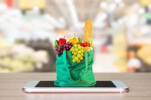 frische lebensmittel und gemüse in grüner einkaufstasche auf mobilem smartphone auf holztisch mit supermarktgang verschwommenem hintergrund lebensmittelgeschäft online-konzept foto
