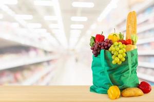 frisches obst und gemüse in wiederverwendbarer grüner einkaufstasche auf holztischplatte mit supermarkt-lebensmittelgeschäft unscharfer defokussierter hintergrund mit bokeh-licht foto