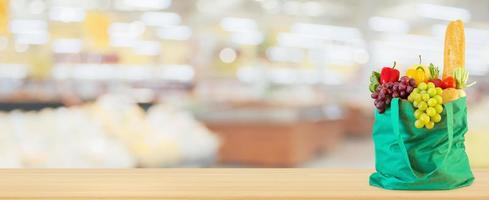 frisches obst und gemüse in wiederverwendbarer grüner einkaufstasche auf holztischplatte mit supermarkt-lebensmittelgeschäft unscharfer defokussierter hintergrund mit bokeh-licht foto