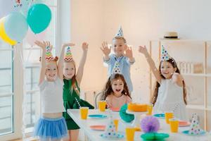 gruppe glücklicher kinder feiern zusammen geburtstag, spielen mit konfetti, tragen partyhüte, posieren in der nähe eines festlichen tisches in einem geschmückten raum mit luftballons, haben überglückliche gesichtsausdrücke, genießen das leben foto