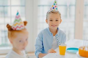 glücklicher kleiner junge trägt partyhut, sitzt am tisch neben dem rothaarigen mädchen, feiert zusammen, feiert geburtstag, posiert in einem großen, geräumigen raum mit fenster. kinder- und urlaubskonzept foto