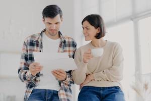 Ernsthaftes Paar studiert Dokumente zusammen, sieht ernst aus, trinkt Kaffee, trägt Freizeitkleidung, plant sein Budget, posiert in einem geräumigen hellen Raum, erledigt Papierkram, bereitet Finanzberichte vor foto