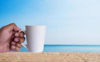 man hand hält weiße peper kaffeetasse zum mitnehmen gelber deckel, gefüllt mit heißem schwarzem kaffee ohne zuckermilch, fertiggetränk, erfrischend. duft wach frisch zur arbeit platziert genießen am meer strand blauer himmel sonnenschein. foto