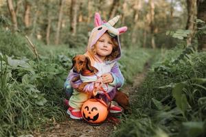 Kleines lachendes Mädchen und ein Zwergdackel in Halloween-Kostümen mit einem Kürbiskorb für Süßigkeiten im Freien. ein Mädchen in einem Regenbogen-Einhorn-Kigurumi-Kostüm, ein Hund in einem Kleid mit vollem Rock. Ansicht von oben foto