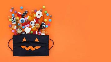 schwarze medizinische maske wie lächelnde kürbis-jack-o-laterne-geschenktüte voller verschütteter traditioneller halloween-süßigkeiten. orangefarbener Bannerhintergrund mit Kopierbereich. foto