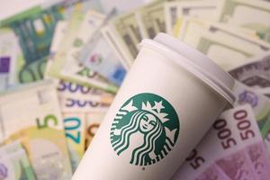charkiw, ukraine - 16. dezember 2021 weißer papierbecher mit starbucks-logo und geldscheinen. Starbucks ist mit über 20.000 Geschäften das größte Kaffeehaus der Welt. foto
