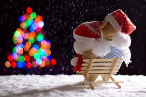 Brauner und weißer Teddybär mit Weihnachtsmütze, der auf einer Holzbank sitzt, wenn es im Winter schneit und bunte Bokeh-Lichter des Weihnachtsbaums betrachtet.
