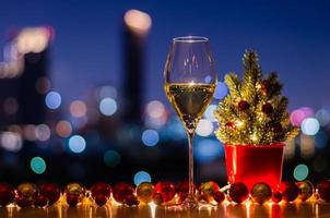 ein glas weißwein, der einen weihnachtsbaum hat, der mit kugelverzierungen und lichtern auf buntem stadt-bokeh-hellhintergrund geschmückt ist. foto