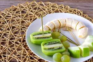veganes Frühstück - Obstvielfalt. äpfel, bananen, kiwi und trauben auf einem teller auf dem grauen holztisch, selektiver fokus, obstsalat foto