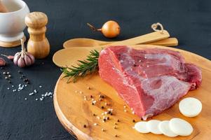 rohes Fleisch Rinderfilet auf Holzschneidebrett, Rosmarinzweige, verschiedene Paprika, Salz und Zwiebel auf dunklem Hintergrund. frisches Rindfleischstück für Steak oder gegrilltes Barbecue
