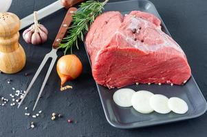 rohes Fleisch Rinderfilet auf Auflaufform, Rosmarinzweige, verschiedene Paprika, Salz und Zwiebel auf dunklem Hintergrund. frisches Rindfleischstück für Steak oder gegrilltes Barbecue foto
