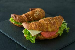 Croissant-Sandwiches mit gesalzenem Lachs auf einem Schreibtisch, serviert mit frischen Salatblättern, Rucola und Gemüse auf schwarzem Hintergrund foto