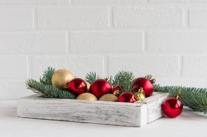 neujahrs- oder weihnachtskomposition. Weiße Holzkiste mit weihnachtlichen roten und goldenen Kugeln, Fichtenzweige auf einem Holztisch gegenüber der Ziegelwand. foto